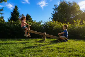 Kinder spielen am Holzzaun auf einer grünen Wiese, umgeben von Bäumen und Sträuchern bei blauem Himmel.