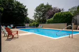 Camping-Pool mit blauem Wasser, Treppe mit Handlauf und zwei Liegestühlen auf der Terrasse neben Bäumen und Hecken.