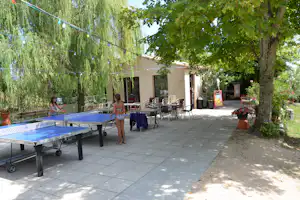 Terrasse am Campingplatz mit Tischtennisplatte, Gartenmöbeln und Lichterkette zwischen Bäumen und einem kleinen Gebäude.