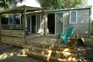 Mobilheim mit Holzterrasse, Stufen und zwei grünen Gartenstühlen vor einer Seitenwand, umgeben von Bäumen.