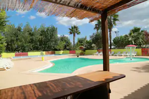 Schwimmbecken am Campingplatz, Blick von der überdachten Terrasse mit Holzdach, Liegen und Palmen im Hintergrund.