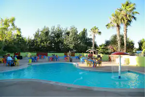 Blauer Camping-Pool mit Springbrunnen, Liegen und Tischen am Beckenrand, Palmen und Bäume im Hintergrund.