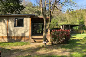 Bungalow auf dem Campingplatz mit Holzterrasse, Sitzbank und Hecke im gepflegten Garten, Bäume und Rasen im Hintergrund.