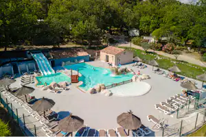 Großes Camping-Schwimmbecken mit Liegen, Sonnenschirmen und Steinterrasse, umgeben von Bäumen und einem kleinen Gebäude.