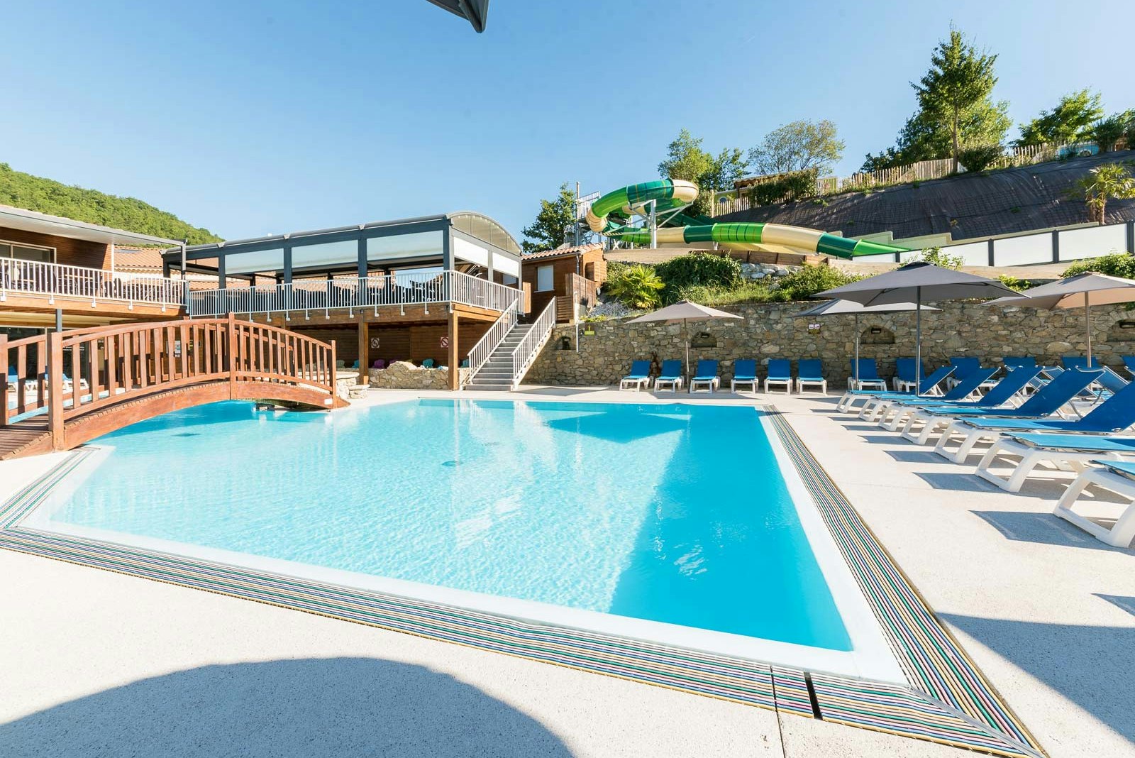 Großer Camping-Pool mit blauen Liegen, Sonnenschirmen und grünen Wasserrutschen neben Holzterrassen und Steinmauer.