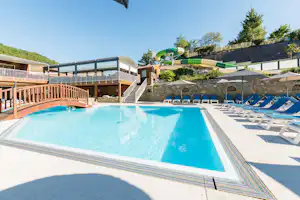 Großer Camping-Pool mit blauen Liegen, Sonnenschirmen und grünen Wasserrutschen neben Holzterrassen und Steinmauer.