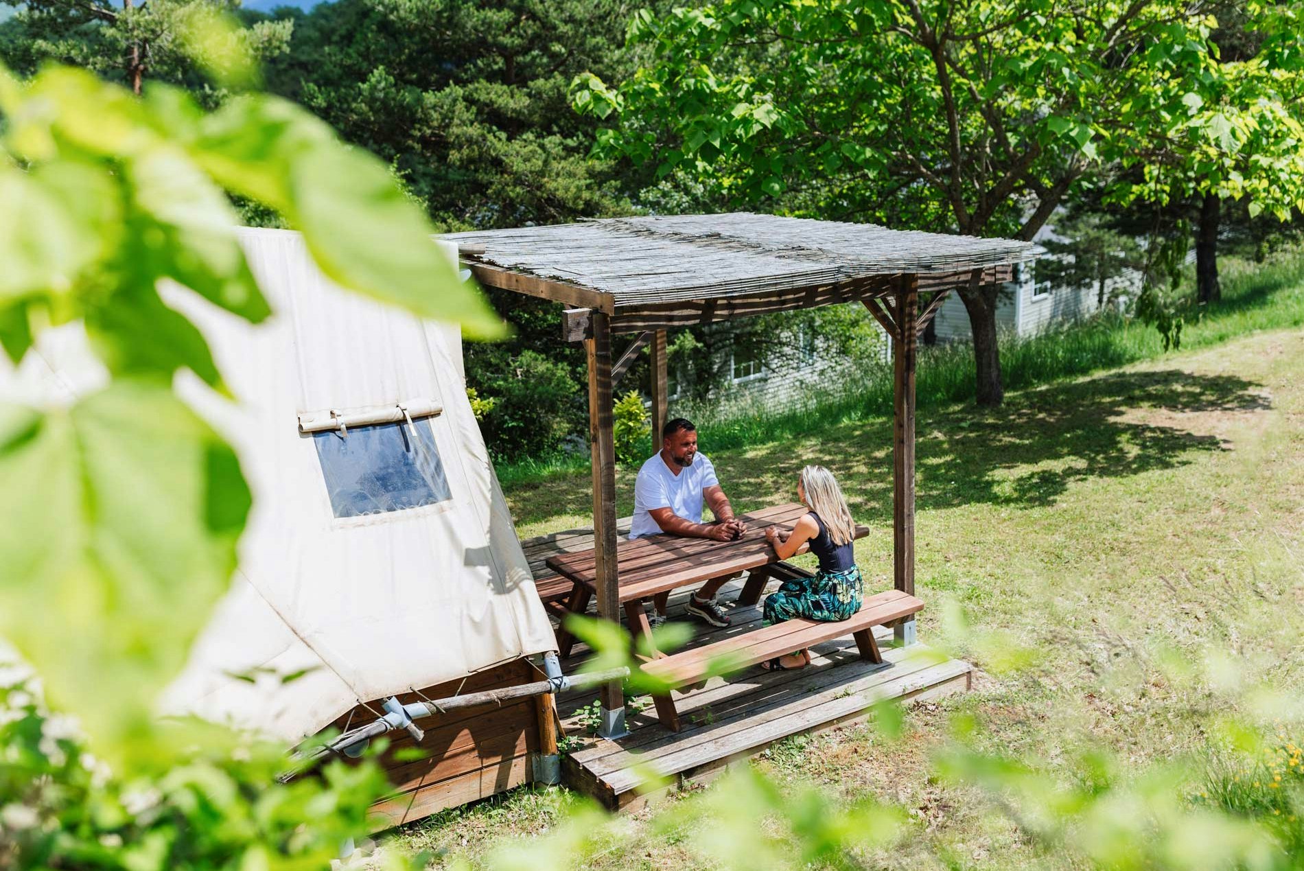 Campingplatz mit Holzterrasse unter einem Baum, zwei Personen am Tisch und Zelt im Vordergrund.
