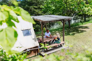 Campingplatz mit Holzterrasse unter einem Baum, zwei Personen am Tisch und Zelt im Vordergrund.