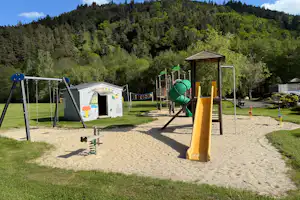 Spielplatz am Campingplatz mit Sandfläche, Rutsche und Klettergerüst, im Hintergrund grüner Wald und Berge.