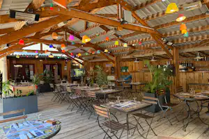 Holzterrasse eines Campingplatzes mit vielen Tischen und Stühlen, Barbereich und bunten Wimpeln unter dem Dach.