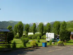 Wohnwagen auf einem Campingplatz mit gepflegtem Garten, Bäumen und Hecken, im Hintergrund Berge und ein Gebäude.