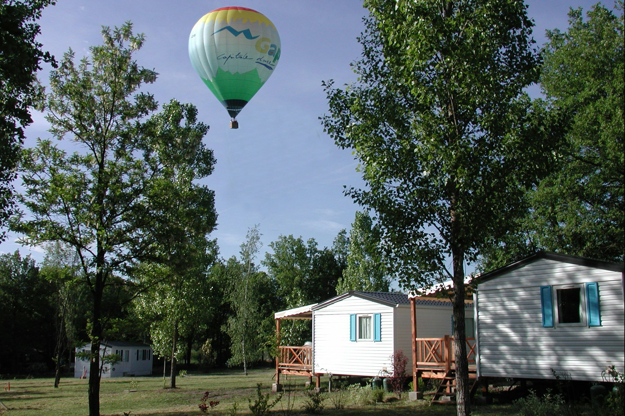 Bunte Heißluftballon am Himmel über einem Campingplatz mit Wohnwagen und Mobilheimen zwischen Bäumen und Rasen.