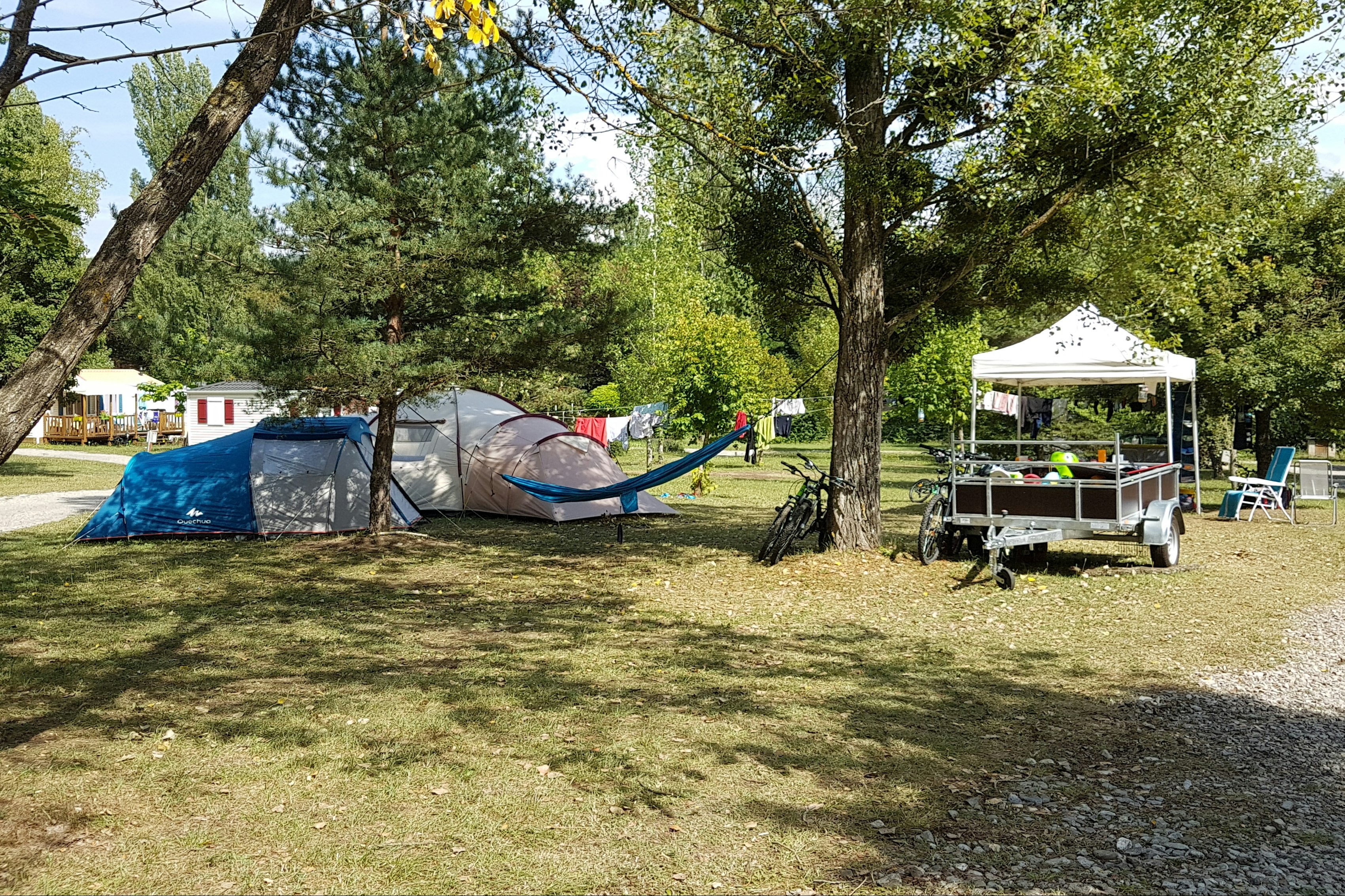 Campingplatz im Grünen: Zelte zwischen Bäumen, daneben ein Grilltisch und ein Pavillon mit Sitzgelegenheiten.