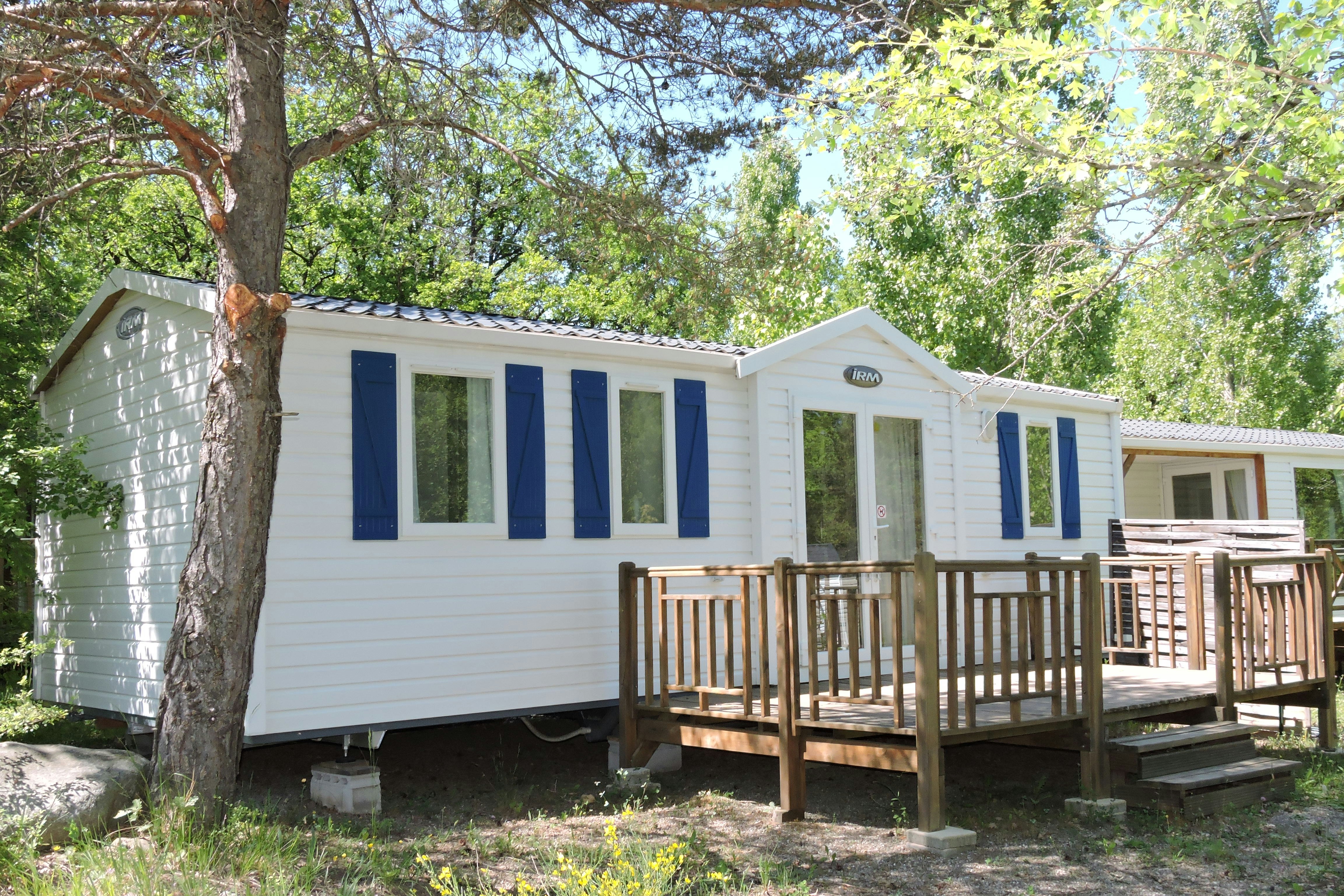 Mobilheim mit Holzveranda und Stufen, weiße Außenwand und blaue Fensterläden, umgeben von Bäumen und Gras.