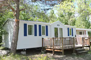 Mobilheim mit Holzveranda und Stufen, weiße Außenwand und blaue Fensterläden, umgeben von Bäumen und Gras.