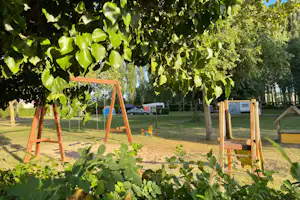 Holzschaukel auf einem Campingplatz, umgeben von Rasen, Bäumen und einem Sandbereich mit Spielzeug.