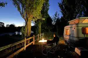 Wohnwagen am Campingplatz am Fluss, davor Campingstühle und Tisch, Laternenlicht zwischen Bäumen bei Abendhimmel.