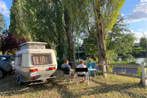 Wohnwagen am See mit Campingstühlen und Tisch unter Bäumen, im Hintergrund grünes Ufer und Landschaft.
