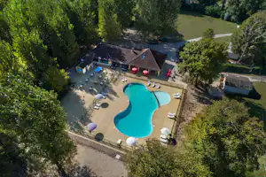 Luftaufnahme vom Campingplatz: Schwimmbecken mit Liegen, Sonnenschirmen und Terrasse am Gebäude, umgeben von Bäumen und Wegen.