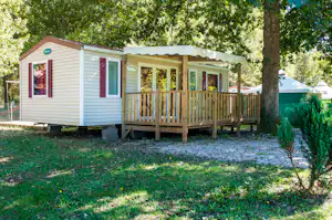 Mobilheim mit Veranda und Holzgeländer im Grünen, umgeben von Bäumen, mit Kiesfläche und kleinen Sträuchern.