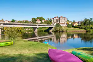 Kanu am Flussufer, Blick auf eine Steinbrücke und Häuser am anderen Ufer, umgeben von Bäumen und Grün.