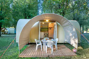 Zelt mit geöffneter Tür, Küchenzeile und Esstisch auf Holzterrasse im Grünen zwischen Bäumen.
