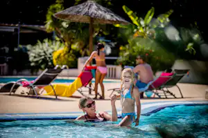 Familie im Campingpool: Kinder planschen im Wasser, im Hintergrund liegen Sonnenliegen und Zelte im Grünen.
