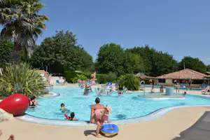 Großes Camping-Schwimmbad mit Palmen, Liegen und Kindern im Wasser, daneben ein Gebäude mit roten Ziegeldächern.
