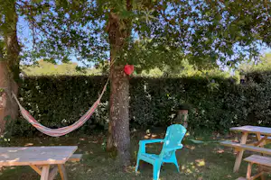 Campinggarten mit Hängematte am Baum, blauem Stuhl und Holztischen auf dem Rasen, Hecke im Hintergrund.