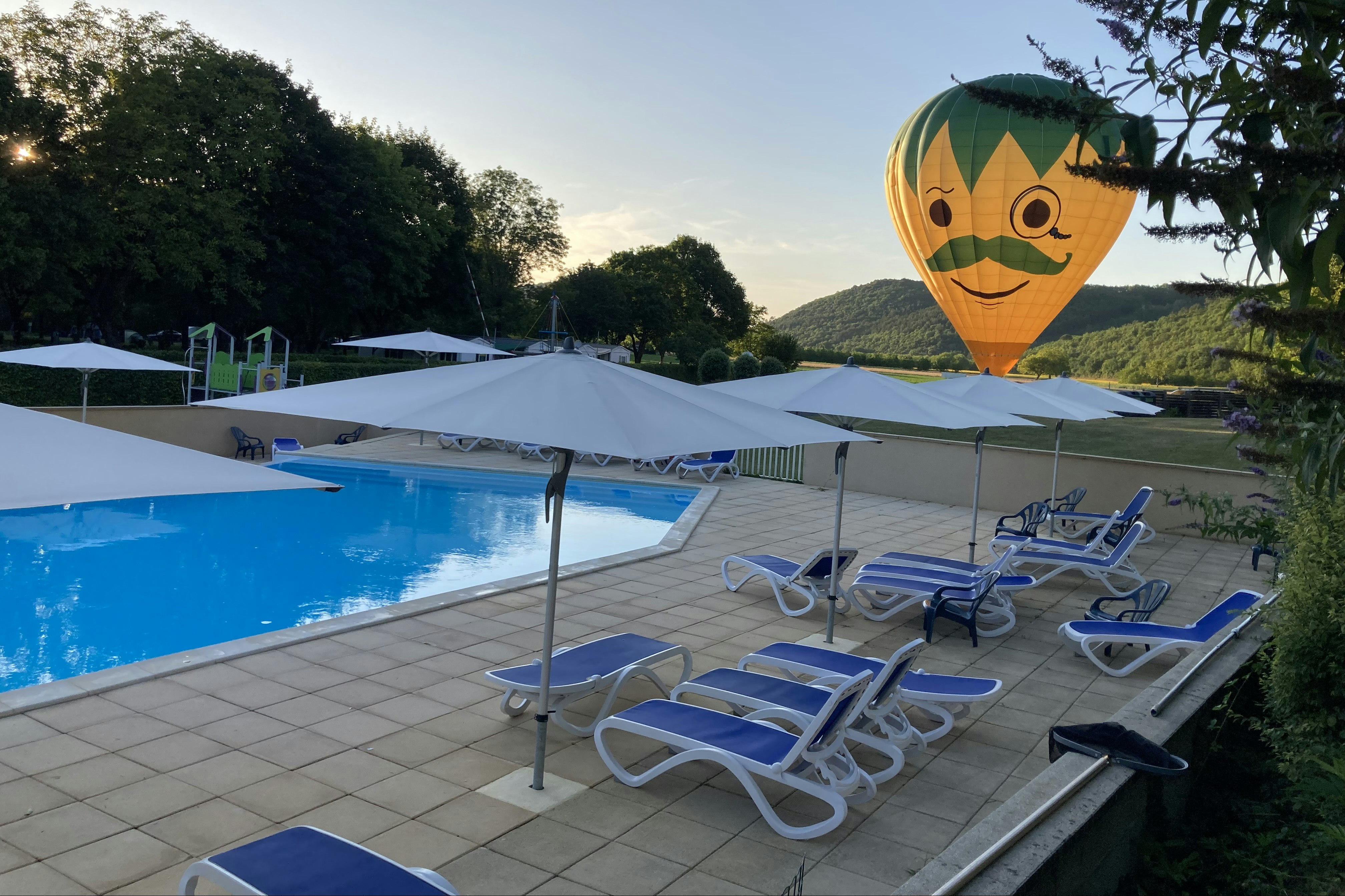 Poolbereich mit mehreren Sonnenliegen und Sonnenschirmen, im Hintergrund ein großer Heißluftballon mit Gesichtsmotiv und grüne Hügel.