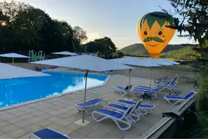Poolbereich mit mehreren Sonnenliegen und Sonnenschirmen, im Hintergrund ein großer Heißluftballon mit Gesichtsmotiv und grüne Hügel.