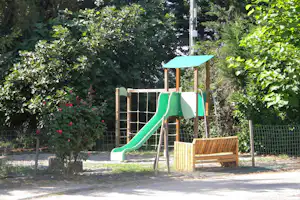 Grüne Rutsche mit Holz-Spielturm und Sonnendach, umgeben von Hecken, Blumen und Zaun im Campinggarten.