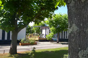 Campingplatz mit Terrasse und Sitzbereich zwischen Bäumen, Holzbänke und Pflanzkübel vor einem Wohnwagen.