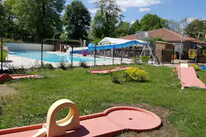 Spielbereich mit Sandspielzeug und Rutsche am Campingplatz, im Hintergrund Pool mit Zaun und Bäumen.