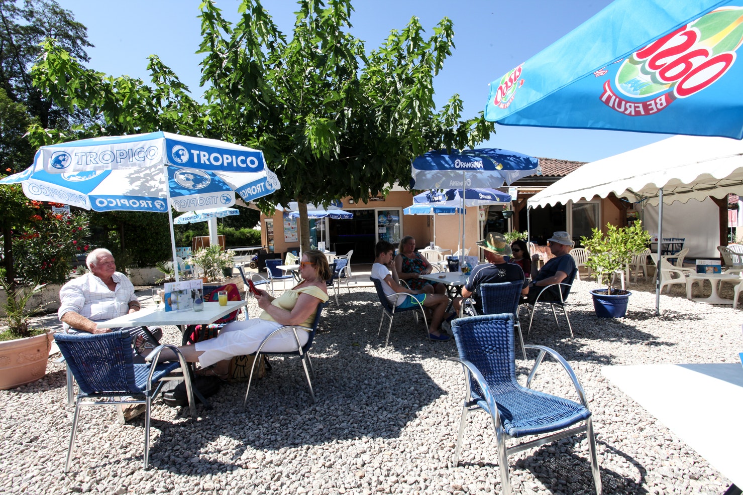 Camping-Terrasse mit blauen Werbeschirmen, Tisch und Stühlen aus Metall, Gäste sitzen im Schatten vor dem Gebäude.