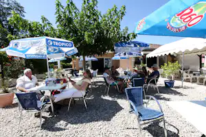 Camping-Terrasse mit blauen Werbeschirmen, Tisch und Stühlen aus Metall, Gäste sitzen im Schatten vor dem Gebäude.