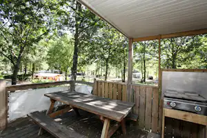 Holz-Terrasse mit Grill und Tisch, dahinter Bäume und Stellplätze mit Wohnmobilen im Campingbereich.