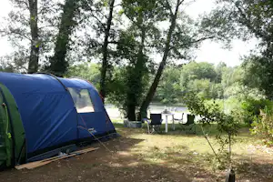Campingzelt am Stellplatz im Grünen, mit Tisch und Stühlen, Bäume und Fluss im Hintergrund.