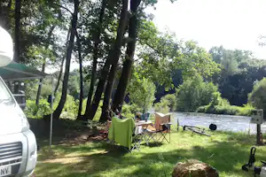 Campingplatz am Fluss mit Wohnwagen, Campingstühlen und Tisch unter großen Bäumen im Grünen.