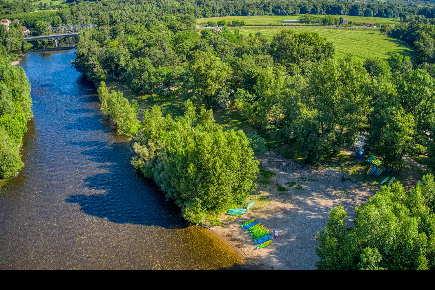 Fluss mit grünem Ufer und Wald, daneben ein Campingbereich mit mehreren bunten Liegen am Ufer.
