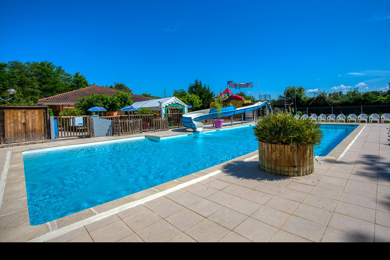 Schwimmbad mit blauem Wasser, Liegen und Sonnenschirmen am Campingplatz, gepflasterte Terrasse, Holzfassade und Bäume im Hintergrund.