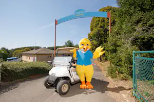 Gelber Kinderfigur am Golfcart vor dem Campingplatz-Eingang, mit Schranke und Hecken entlang des Weges.