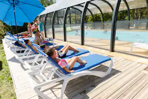 Sonnenliegen am Pool unter einer Glasüberdachung, daneben blaue Sonnenschirme und Holzterrasse auf dem Campingplatz.
