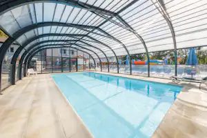 Überdachtes Schwimmbecken mit blauem Wasser, Liegen und Glaswänden im Poolbereich, dahinter Gebäude und Zaun.