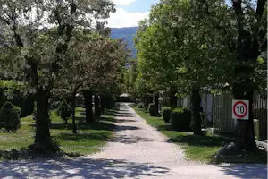 Schotterweg auf einem Campingplatz zwischen Bäumen, rechts ein Zaun und ein Schild mit 10, im Hintergrund Berge.