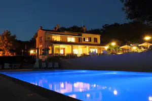 Beleuchtetes Camping-Gebäude mit Pool am Abend, Liegestühle und Zaun im Vordergrund, Bäume im Hintergrund.