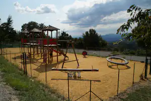 Spielplatz auf dem Campinggelände: Sandfläche, Rutsche, Klettergerüst und Wippe, umgeben von Zäunen und Bäumen.