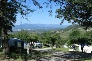 Campingplatz unter Bäumen mit Wohnwagen, Zelten und Autos am Weg, dahinter weite grüne Hügellandschaft und Berge.