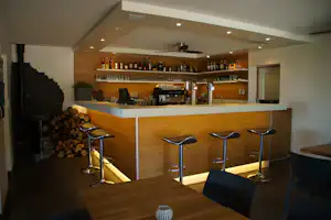 Campingbar mit Holztheke, Getränkeflaschen im Regal, Barhockern und Kamin mit Holzscheiten im Hintergrund.