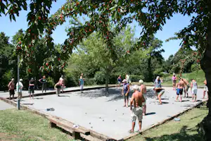 Petanque-Spiel auf einem Betonplatz im Grünen, umgeben von Bäumen mit roten Früchten und mehreren Spielern.
